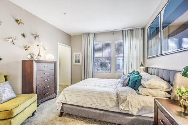 Bedroom with plush bed, dresser with decor, large framed artwork, and large windows.