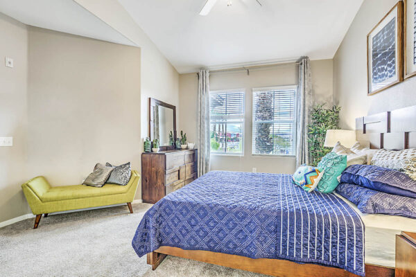 Bedroom with plush bed, daybed with pillows, dresser with mirror, and large windows.