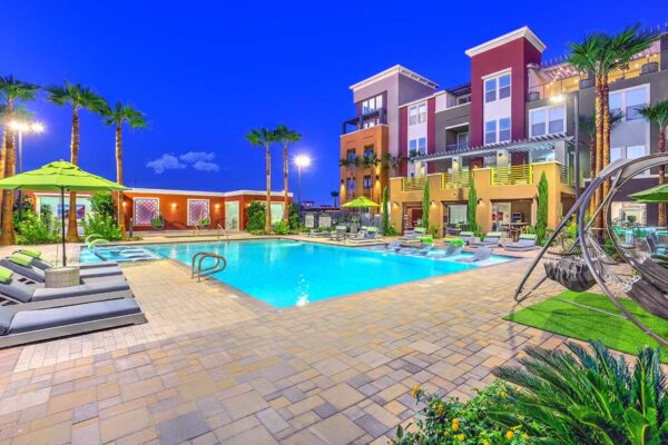 Pool area with stone paver patio, lounge chairs, umbrellas, and gazebos overlooked by apartment balconies.