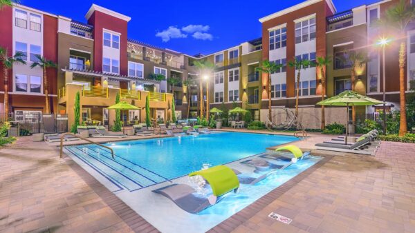 Pool area with stone paver patio, lounge chairs, umbrellas, and gazebos overlooked by apartment balconies.