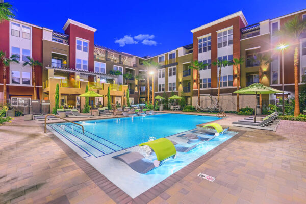 Pool area with stone paver patio, lounge chairs, umbrellas, and gazebos overlooked by apartment balconies.