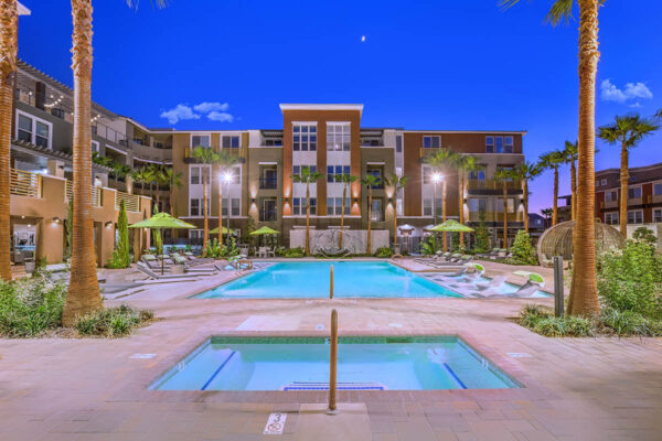 Pool area with stone paver patio, lounge chairs, umbrellas, and gazebos overlooked by apartment balconies.