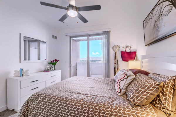 Bedroom with plush bed, dresser with decor, framed mirror, ceiling fan, and door to balcony.