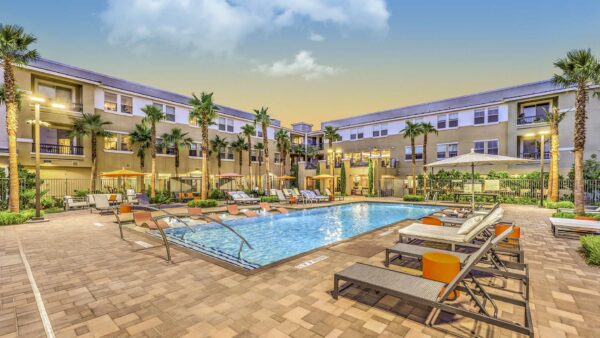 Pool area with lounge chairs, tables with umbrellas, and tall palm trees.