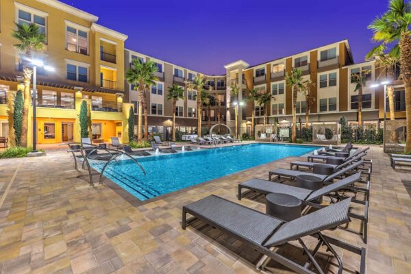 Pool area with stone paver patio, lounge chairs, lush landscaping, and tall palm trees.