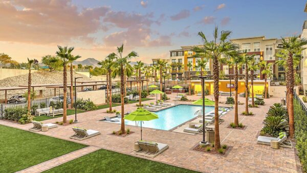 Pool area with stone paver patio, lounge chairs, umbrellas, lush landscaping, and palm trees.
