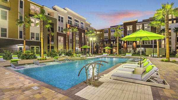 Pool area with stone paver patio, lounge chairs, umbrellas, lush landscaping, and palm trees.