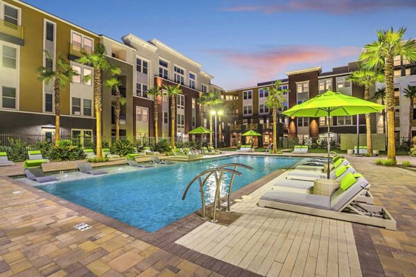 Pool area with stone paver patio, lounge chairs, umbrellas, lush landscaping, and palm trees.