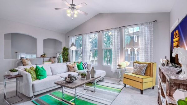 Living room with carpet, plush seating, ceiling fan, shelves with decor, and large windows.