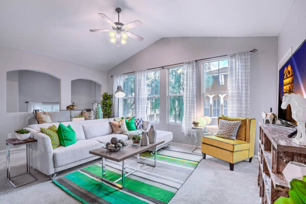 Living room with carpet, plush seating, ceiling fan, shelves with decor, and large windows.