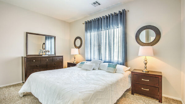 Bedroom with plush bed, bedside dressers with lamps, dresser with mirror, and large window.