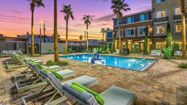 Pool area with stone paver patio, lounge chairs, and tall palm trees overlooked by apartments.
