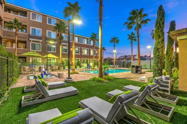Pool area with stone paver patio, lounge chairs, and tall palm trees overlooked by apartments.