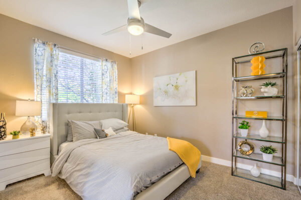 Bedroom with carpet, plush bed with headboard, shelves with decor, ceiling fan, and large window.