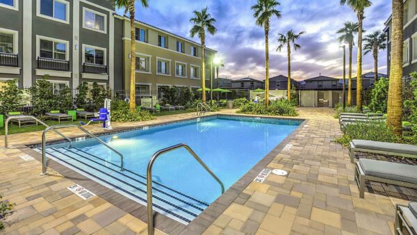 Pool area with stone paver patio, lounge chairs, lush landscaping, and tall palm trees.