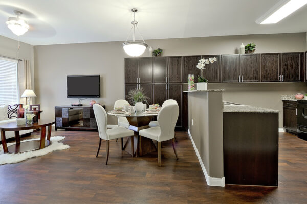 Apartment kitchen with wood floor and cabinets, stone counters, black appliances, and dining table with chairs.