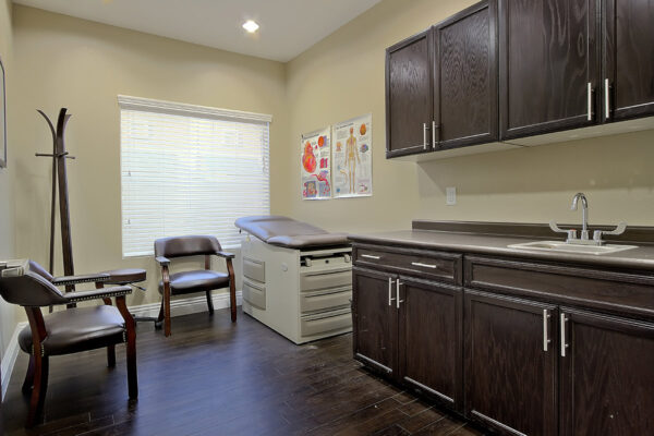 Exam room with wood floor and cabinets, plush chairs, exam table, and large window.