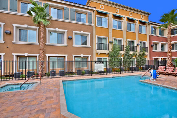 Pool area with stone paver patio, lounge chairs, lush landscaping, and tall palm trees.