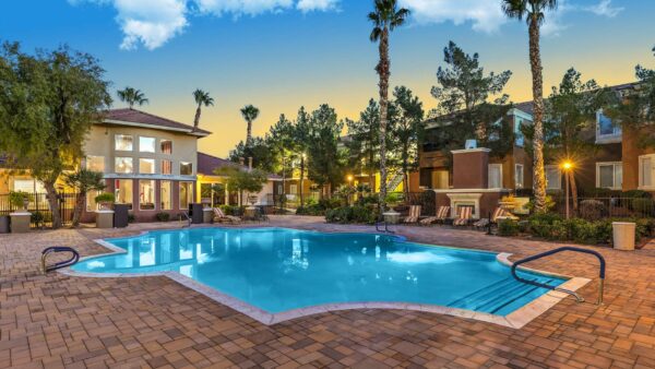 Pool area with stone paver patio, lounge chairs, lush landscaping, and tall palm trees.