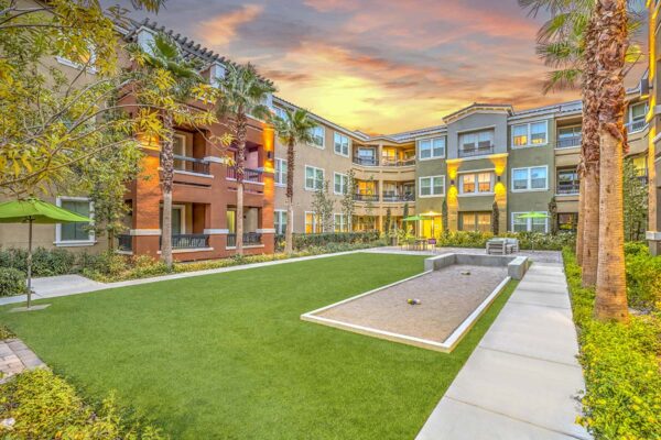 Courtyard with bocce ball court, grassy lawn, umbrellas, lush landscaping, and tall palm trees.