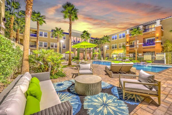 Pool area with stone paver patio, plush seating, tables with umbrellas, and tall palm trees.