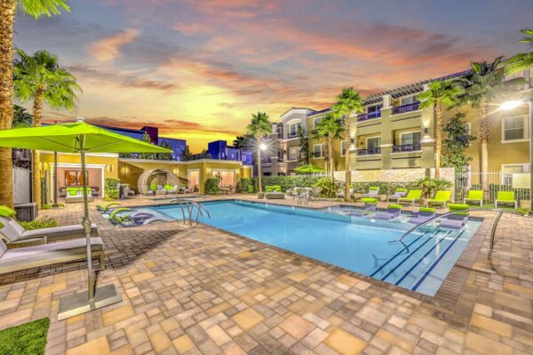 Pool area with stone paver patio, plush seating, tables with umbrellas, and tall palm trees.