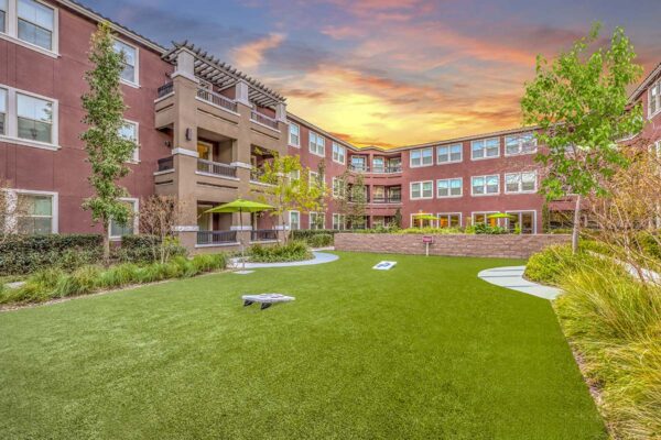 Courtyard with grassy lawn, bag toss game, lush landscaping, and tall trees.