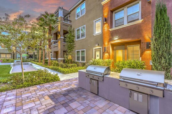 Outdoor grilling area with stone paver patio, built in BBQ grills, lush landscaping, and tall trees.