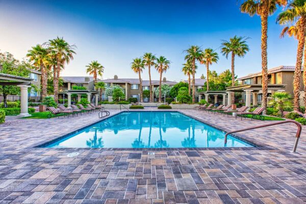 Pool area with stone paver patio, lounge chairs, lush landscaping, and tall palms.