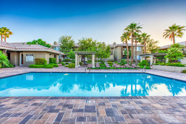 Pool area with stone paver patio, lounge chairs, lush landscaping, and tall palms.
