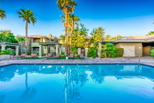 Pool area with stone paver patio, lounge chairs, lush landscaping, and tall palms.