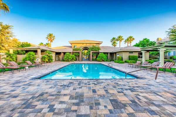 Pool area with stone paver patio, lounge chairs, lush landscaping, and tall palms.