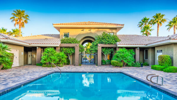 Pool area with stone paver patio, clubhouse, lush landscaping, and tall palms.