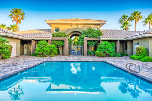 Pool area with stone paver patio, clubhouse, lush landscaping, and tall palms.