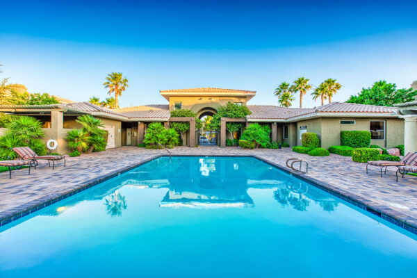 Pool area with stone paver patio, lounge chairs, lush landscaping, and tall palms.