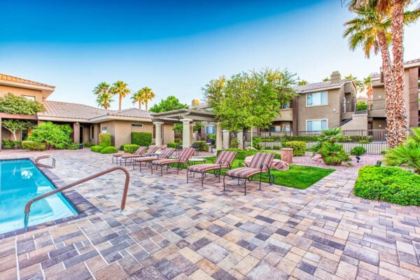 Pool area with stone paver patio, lounge chairs, lush landscaping, and tall palm trees.