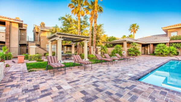 Pool area with stone paver patio, lounge chairs, lush landscaping, and tall palm trees.
