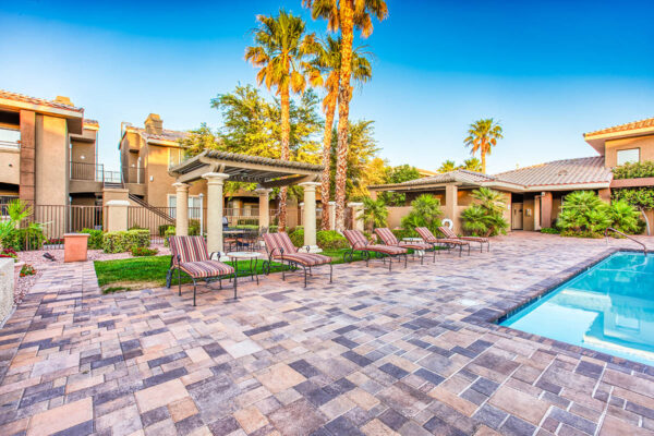 Pool area with stone paver patio, lounge chairs, lush landscaping, and tall palm trees.