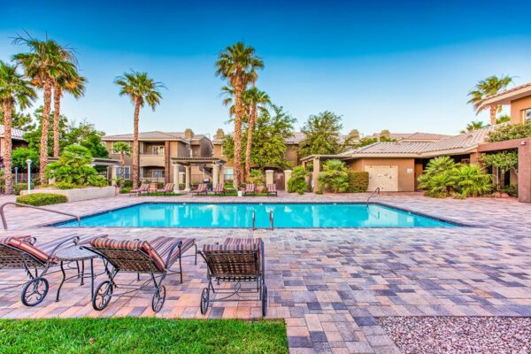 Pool area with stone paver patio, lounge chairs, lush landscaping, and tall palm trees.