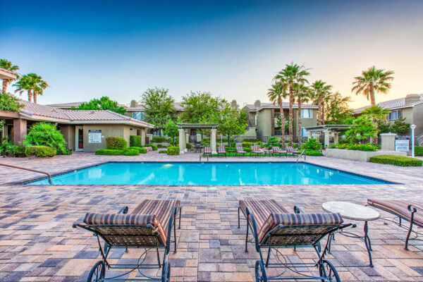 Pool area with stone paver patio, lounge chairs, lush landscaping, and tall palm trees.