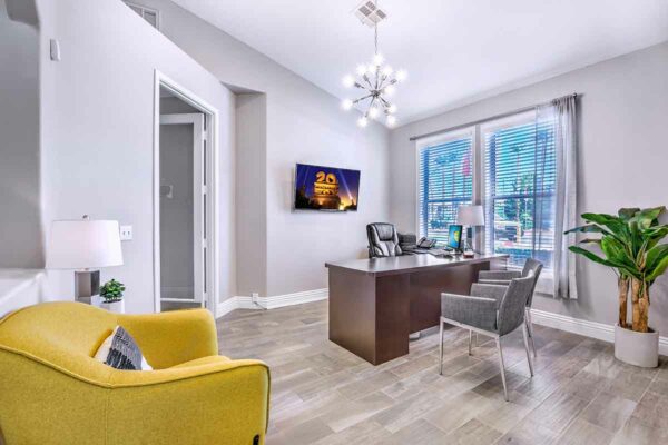 Office with computer desk, plush chairs, bright chandelier, and large window.