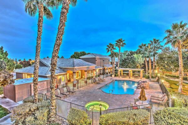 Fenced pool area with lounge chairs, pergola, rocky waterfall, and tall palm trees.