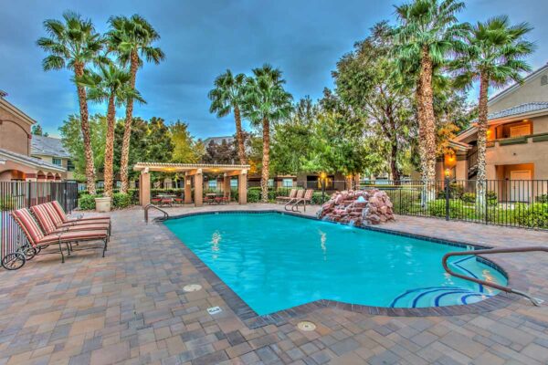 Swimming pool with rock waterfall, lounge chairs, metal fence, and tall palm trees.