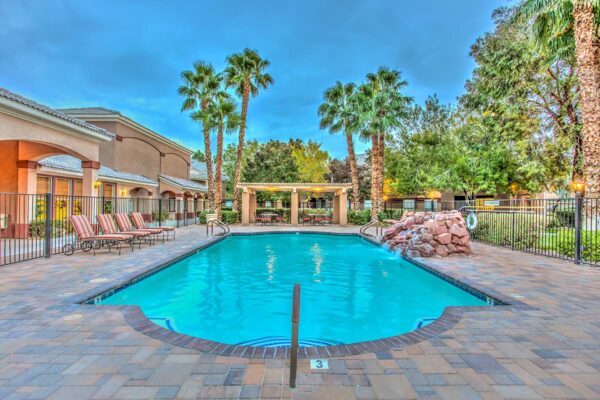 Swimming pool with rock waterfall, lounge chairs, metal fence, and tall palm trees.
