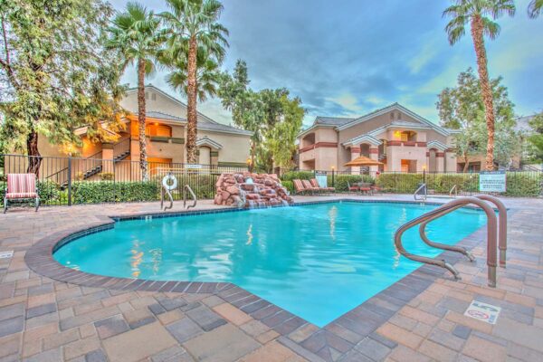 Swimming pool with rock waterfall, lounge chairs, metal fence, and tall palm trees.