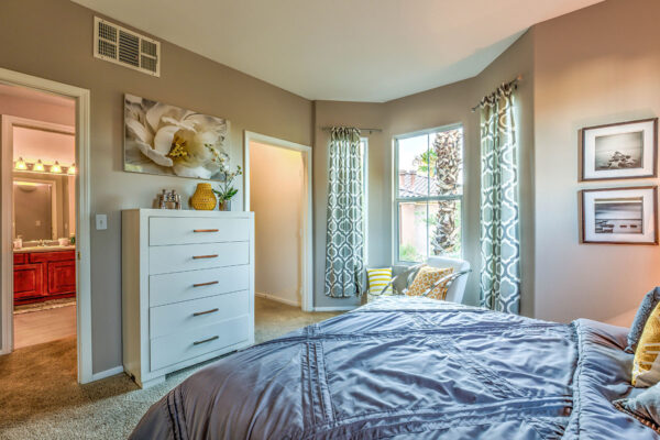 Bedroom with carpet, plush bed with headboard, dresser with decor, and large windows.