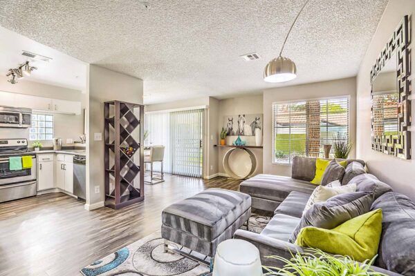 Living room with plush furniture, shelves with decor and wine, modern kitchen, and balcony door.