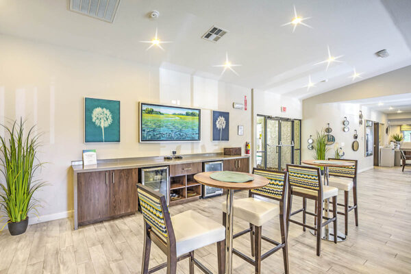 Clubroom with tall bar tables and chairs, demo kitchen with fridges, and framed artwork.