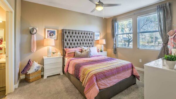 Bedroom with carpet, plush bed with headboard, drawers with lamps, ceiling fan, and large windows.