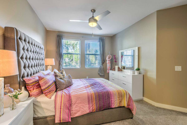 large one-bedroom in Summerlin apartment with carpet, plush bed with headboard, drawers with lamps, ceiling fan, and large windows.